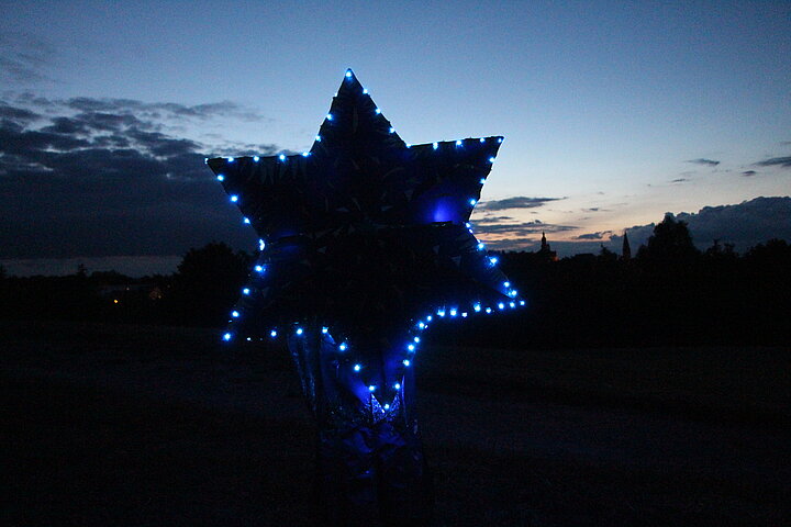 Fotoaufnahme der Museumsnacht. Im Fokus steht ein sechseckiger Stern, an dessen Rändern kleinen Lämpchen leuchten. Im Hintergrund sind der dunkle Himmel und die Landschaft erkennbar. Die Lämpchen am Stern sind die einzige Lichtquelle. 